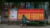 中国新疆叶尔羌地区的一名妇女坐在公交车站，车站张贴政府的宣传标语：“建设美丽新疆，共圆祖国梦想”。(2023年7月17日)