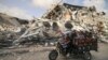 Një burrë palestinez transporton fëmijët ndërsa në sfond duket një godinë e shkatërruar nga sulmet ajrore izraelite (21 maj 2021)