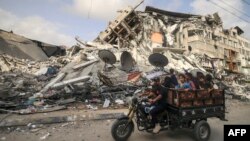 Një burrë palestinez transporton fëmijët ndërsa në sfond duket një godinë e shkatërruar nga sulmet ajrore izraelite (21 maj 2021)